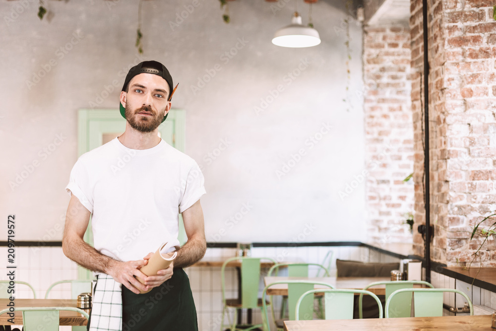 Young waiter in black cap,white t-shirt and apron standing with notepad in hands and dreamily looking in camera while working in cafe