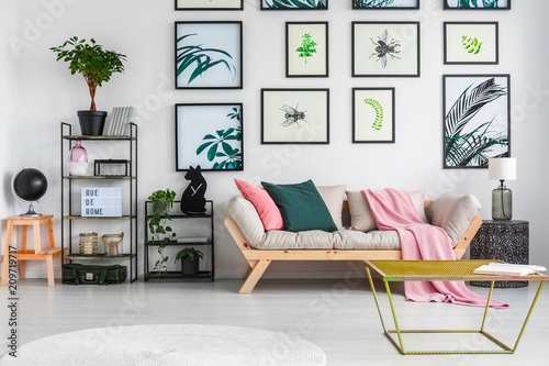 Gold metal table standing in white living room interior with decor on black metal racks, fresh green plants, light grey sofa with cushions and pink blanket and posters hanging on the wall