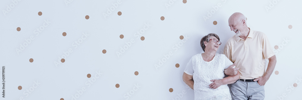 Happy, elderly couple in love looking at each other while posing on a white wall with polka dot pattern and copy space. Happy, elderly couple in love looking at each other while posing on a white wall with polka dot pattern and copy space.