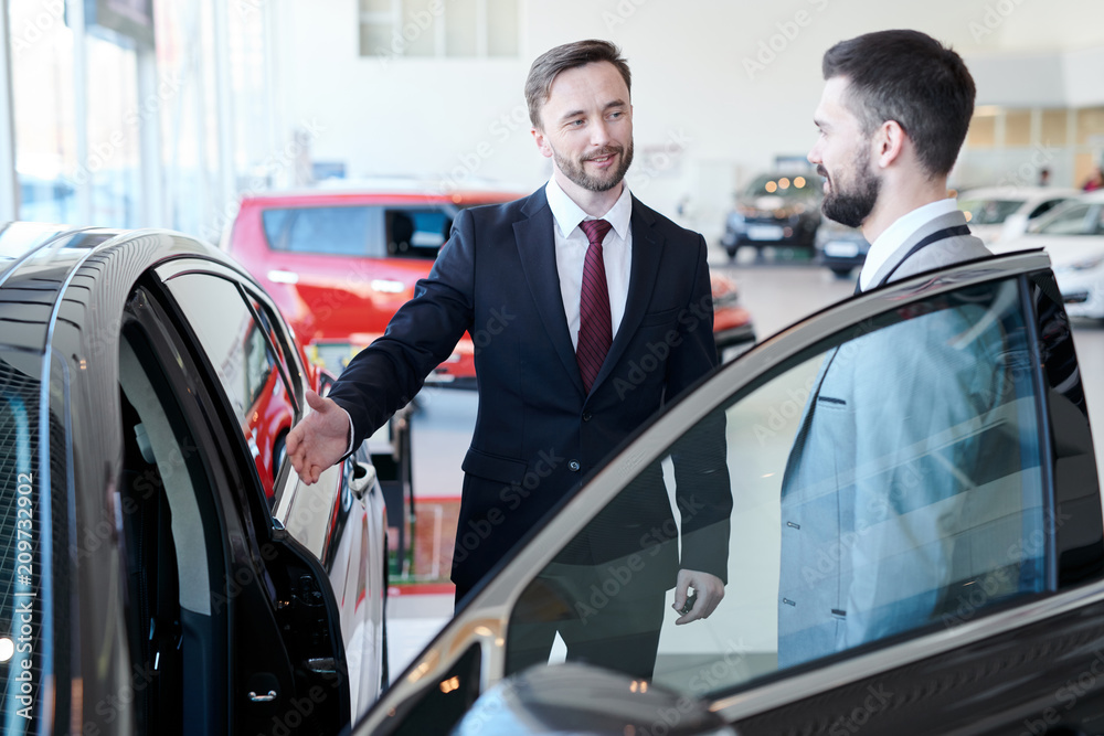 Portrait of handsome car salesman showing brand new luxury car to smiling client standing ion luxury dealership, copy space Portrait of handsome car salesman showing brand new luxury car to smiling client standing ion luxury dealership, copy space