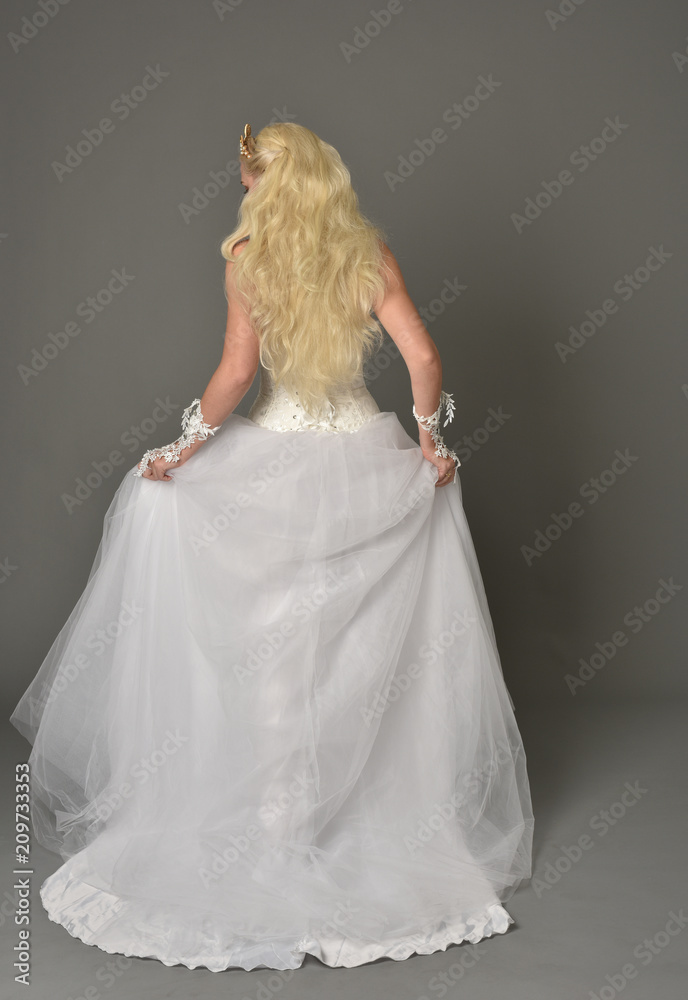 full length portrait of blonde girl wearing white gown, standing pose with back to the camera. grey studio background.