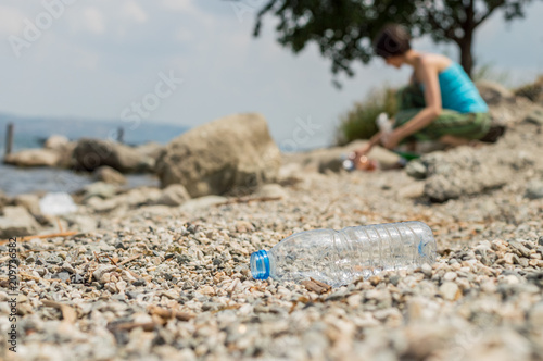 Plastic bottle dumped on the coastal strip with blurry woman picking up the garbage on the background.