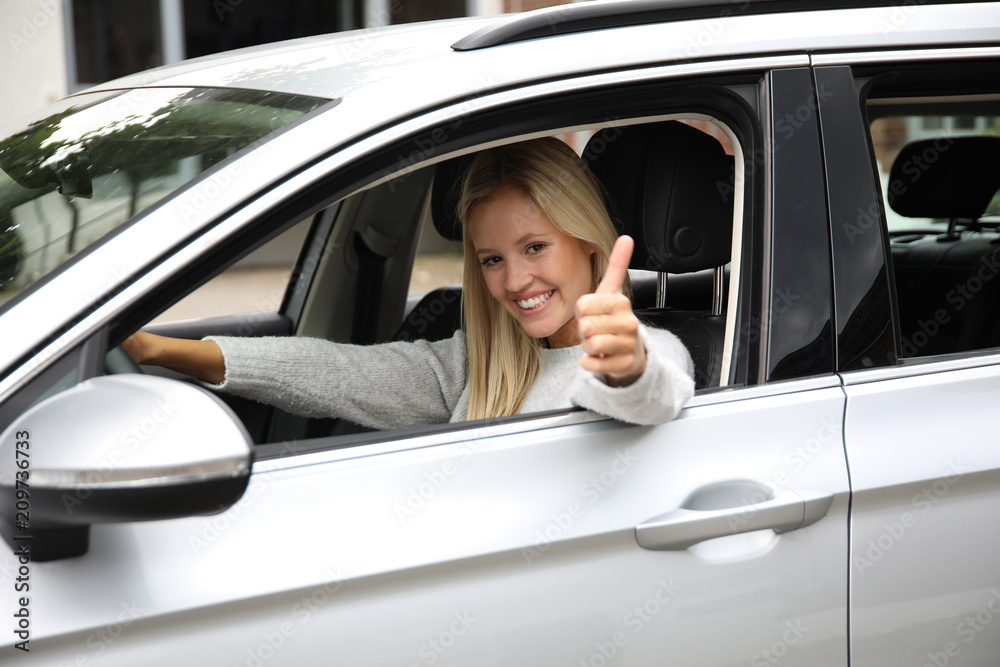 Hübsche blonde Frau sitzt am Lenkrad eines Autos und zeigt lachend Daumen hoch