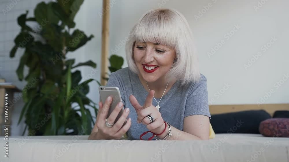 The girl smiles when watching a photo on her phone. A woman in the bed on the background of a white wall and a large bowl is fun looking at her smartphone. The girl smiles when watching a photo on her phone. A woman in the bed on the background of a white wall and a large bowl is fun looking at her smartphone.