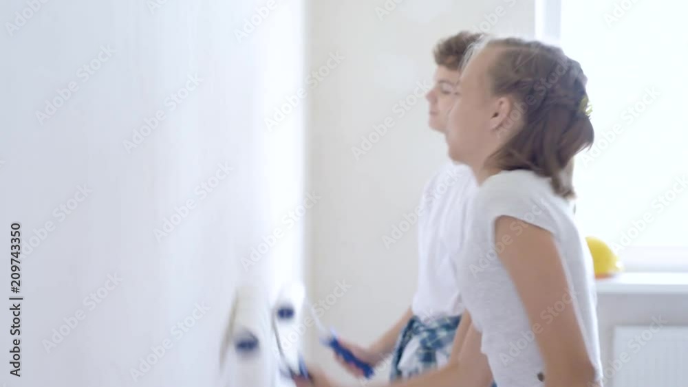 Teen boy and girl makes repairs in new flat. Sister and brother painting wall in apartment room. Children paints the wall, using a rollers. Young friends laughing and having fun. 