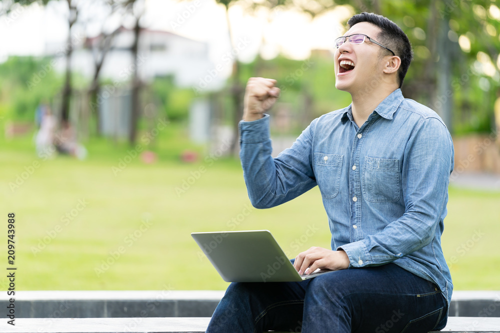 Attractive asian happy man gesture or raise hand excited screaming yes reading online good news or social network in laptop sitting in city park with copy space. Winner or victory expression concept. Attractive asian happy man gesture or raise hand excited screaming yes reading online good news or social network in laptop sitting in city park with copy space. Winner or victory expression concept.