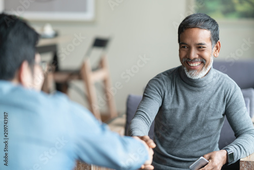 Confident mature Asian man sitting, smiling and shaking hand with partnership after making profitable agreement. Smart man handshake, deal and greeting concept.