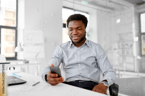 business, technology and people concept - happy african american businessman with headphones and smartphone listening to music at office