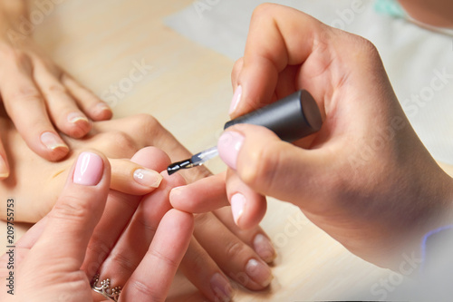 Manicurist applying colorless varnish. Female hands applying transparent nail polish on healthy nails of young woman. Manicurist making manicure.