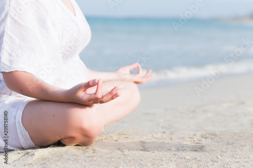 Young beautiful pregnant woman in white dress doing yoga exercises on the beach, touching her belly, making heart shape, breathing and relax with love and care at calm blue sea on white sand, close-up