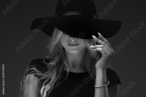 elegant slender girl model in a stylish narrow black dress and a wide hat on a black background. She holds his fingers over his hat, only his lips can be seen photo