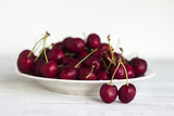 Ripe cherry berries with water droplets on a white plate on a white wooden background, clean, healthy and tasty fruit.