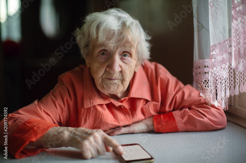Old woman takes on smartphone sitting at the table in the house.