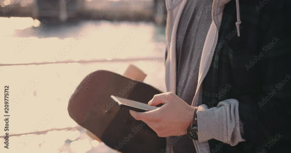 Hipster teenage skateboarder texting and walking on jetty
