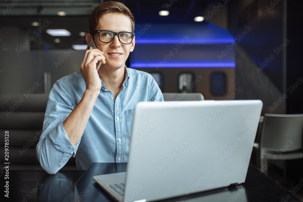 Portrait of a young man with a good mood, a businessman in a shirt and glasses, who works on a laptop and talking on the phone in a cafe, can be used for advertising, text insertion. Portrait of a young man with a good mood, a businessman in a shirt and glasses, who works on a laptop and talking on the phone in a cafe, can be used for advertising, text insertion.