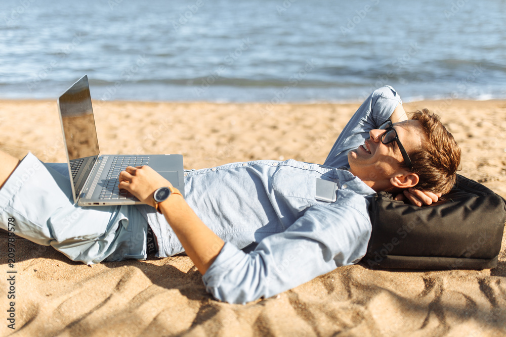 Young guy with glasses, lying on the sand, working on his laptop on the beach, against the sea, working on vacation, suitable for advertising, text insertion