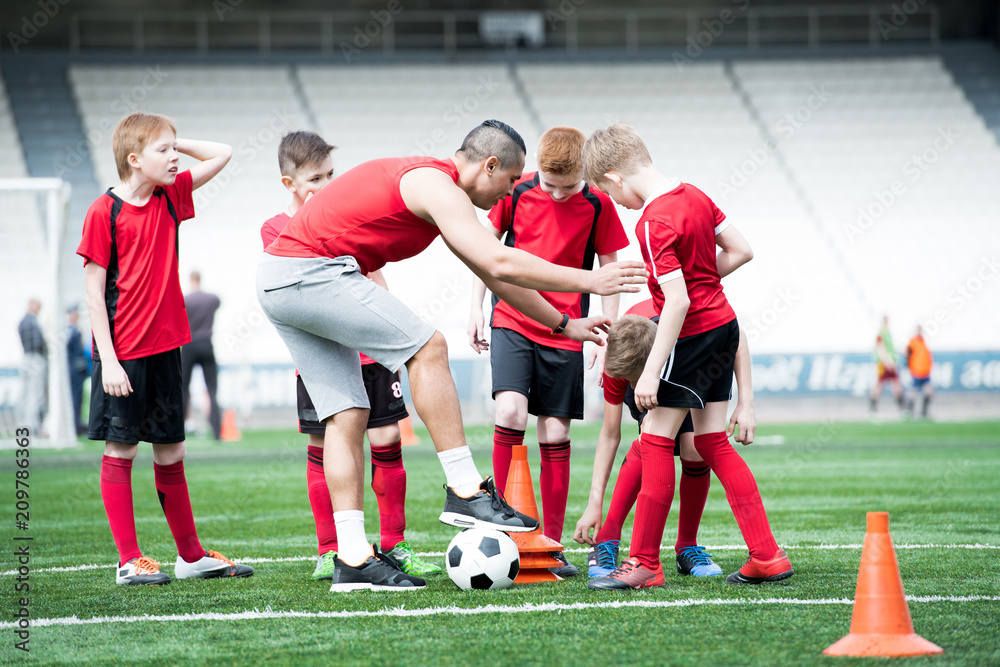 Full length portrait of junior football team practicing in stadium, focus on coach giving instructions to boys, copy space