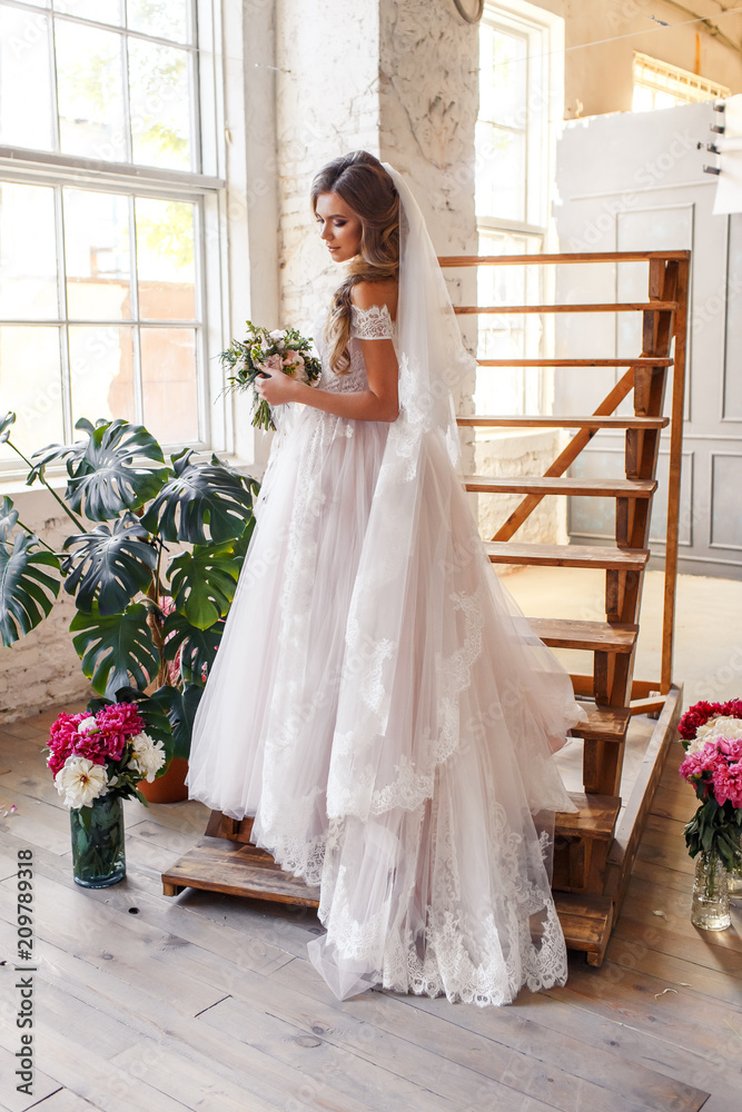 Fototapeta premium A bride with hairstyle and make up in gorgeous pink wedding dress and a vail with a bouquet of peonies. A portrait of beautiful girl with brunette hair and blue eyes in studio