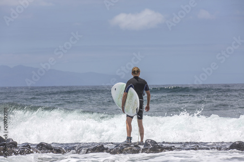 The surf enters the water. Male surfer entering the sea with his board in a black surfing suit. Tenerife, Spain. Surfer entering the ocean .Ready for a great surfing day.