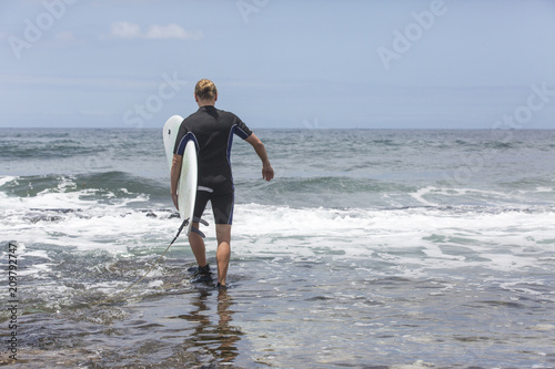 The surf enters the water. Male surfer entering the sea with his board in a black surfing suit. Tenerife, Spain. Surfer entering the ocean .Ready for a great surfing day.