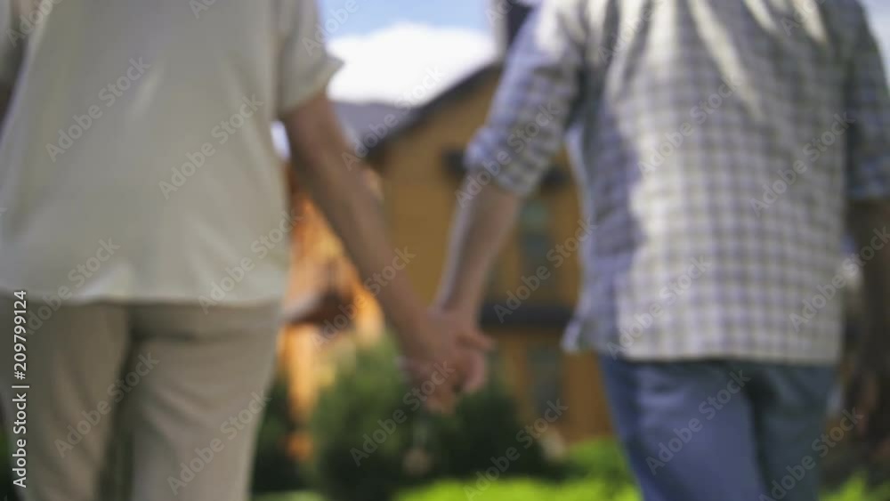 Close-up of elderly couple's hands holding house door key and walking together on frontyard green lawn towards their new big dream house. Happy senor couple retired and purchased new home Close-up of elderly couple's hands holding house door key and walking together on frontyard green lawn towards their new big dream house. Happy senor couple retired and purchased new home