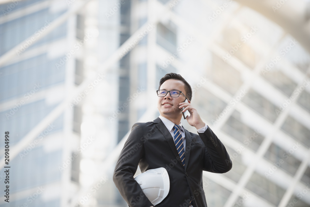 Portrait of a happy young asian businessman talking on mobile phone and holding a helmet. Man outdoor with phone in a city Bangkok Thailand. Concept of communication. Portrait of a happy young asian businessman talking on mobile phone and holding a helmet. Man outdoor with phone in a city Bangkok Thailand. Concept of communication.