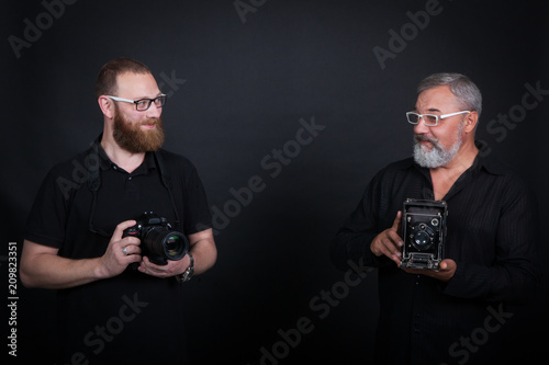 Two male photographers, young and elderly, with cameras - modern digital and vintage widescreen. On a black background in the studio.