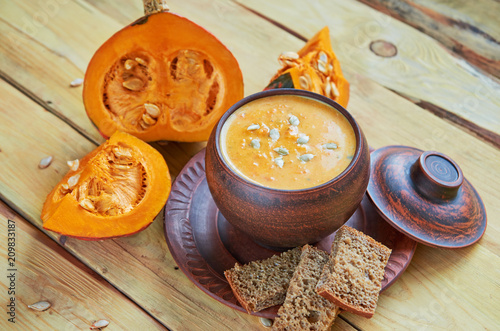 Bowl of spicy pumpkin soup with sliced yellow pumpkins, seeds and bread on the wooden background. Traditional autumn food or thanksgiving dish. Side view