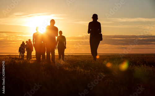 A group of people walking in a track. They go against the background of the orange sun, their contours and silhouettes are visible.
