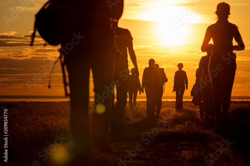 A group of people walking in a track. They go against the background of the orange sun, their contours and silhouettes are visible.

