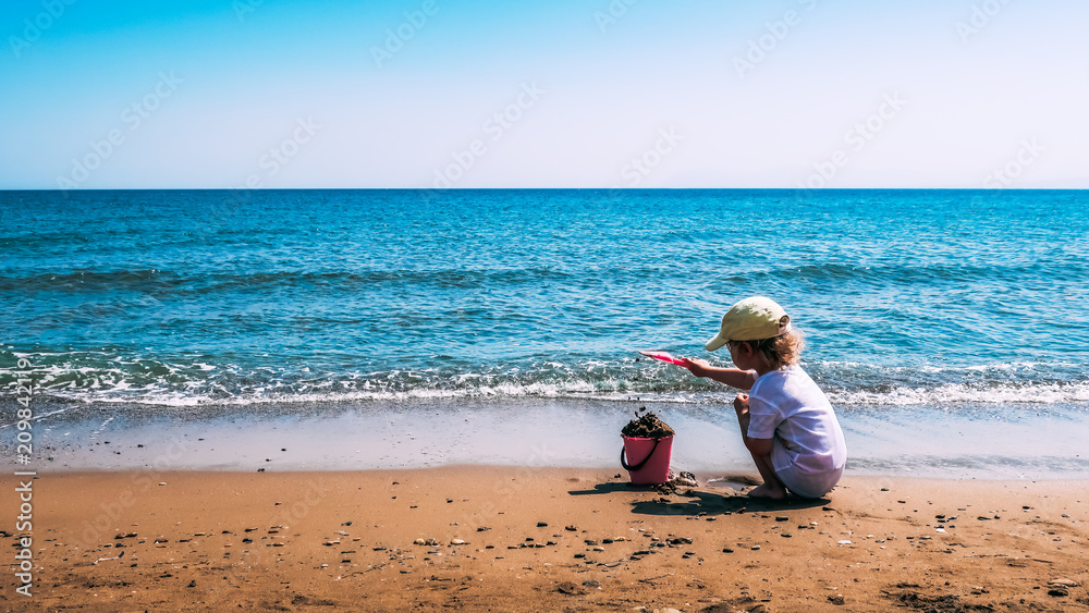 A child plays with a pink plastic bucket and shovel in the sand on a beach, close to the sea. Crete, Greece. A child plays with a pink plastic bucket and shovel in the sand on a beach, close to the sea. Crete, Greece.