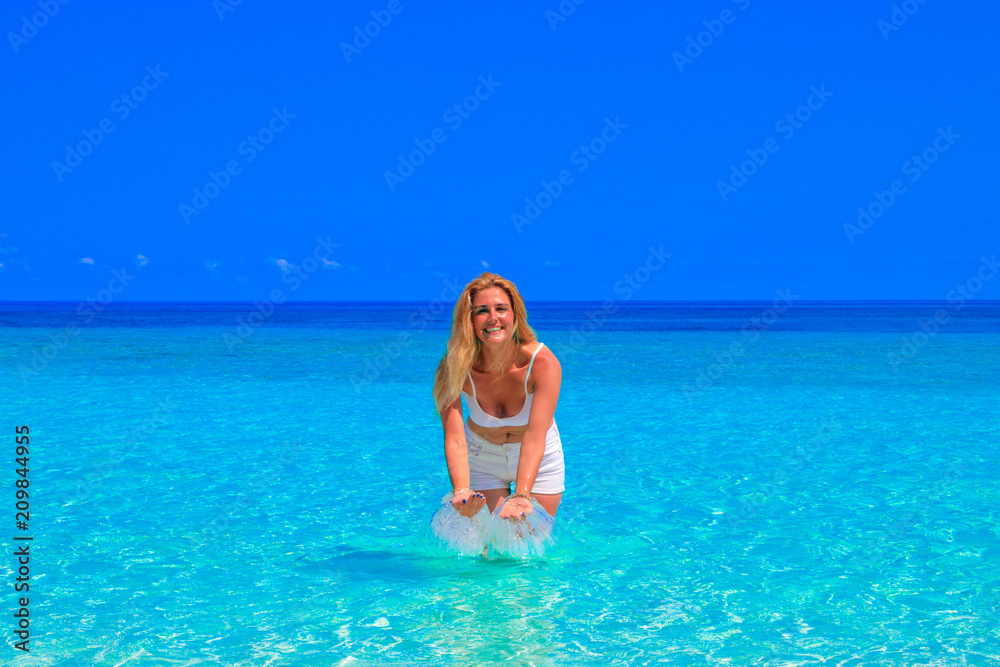 Gorgeous blond woman with white clothes is standing in the water at the sandy beach