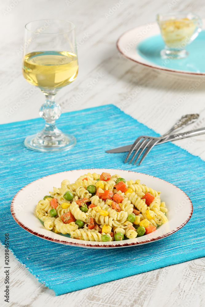 Delicious homemade fusilli pasta cooked with basil pesto, carrots, corn and peas in a plate on a turquoise blue napkin, served with glass of white wine and mustard-mayonnaise sauce