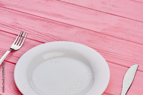 Empty plate, fork and knife on wood. Pink wooden table setting with white plate and stainless fork and knife, copy space.