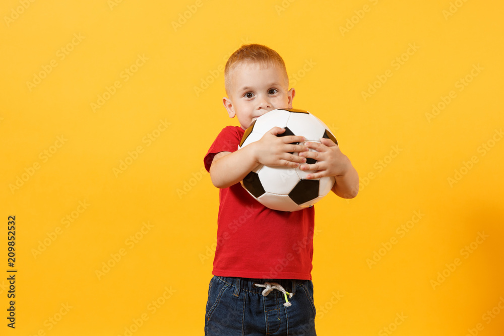 Little cute kid baby boy 3-4 years old, football fan in red t-shirt holding in hand soccer ball isolated on yellow background. Kids sport family leisure lifestyle concept. Copy space for advertisement Little cute kid baby boy 3-4 years old, football fan in red t-shirt holding in hand soccer ball isolated on yellow background. Kids sport family leisure lifestyle concept. Copy space for advertisement