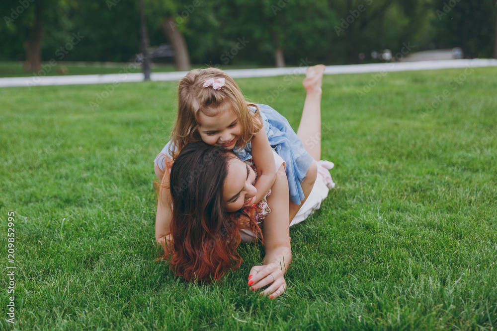 Fototapeta premium Joyful woman in light dress and little cute child baby girl lie on green grass in park rest play and have fun. Mother, little kid daughter. Mother's Day, love family, parenthood, childhood concept.