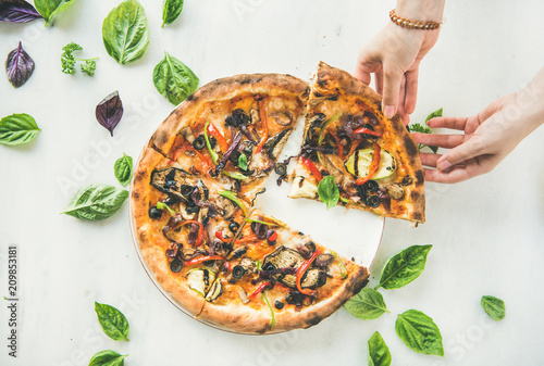 Summer dinner or lunch. Flat-lay of female hands taking freshly baked Italian vegetarian pizza with vegetables and fresh basil over white marble table, top view