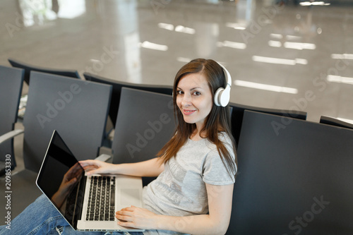 Young smiling traveler tourist woman with headphones listen music working on laptop with blank screen wait in lobby hall at airport. Passenger traveling abroad on weekends getaway. Air flight concept.