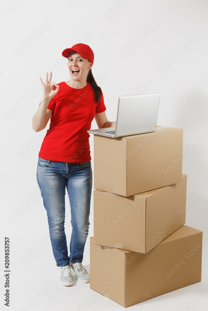 Full length portrait of delivery young woman in red cap, t-shirt isolated on white background. Female courier standing near empty cardboard boxes with laptop pc computer. Receiving package. Copy space Full length portrait of delivery young woman in red cap, t-shirt isolated on white background. Female courier standing near empty cardboard boxes with laptop pc computer. Receiving package. Copy space