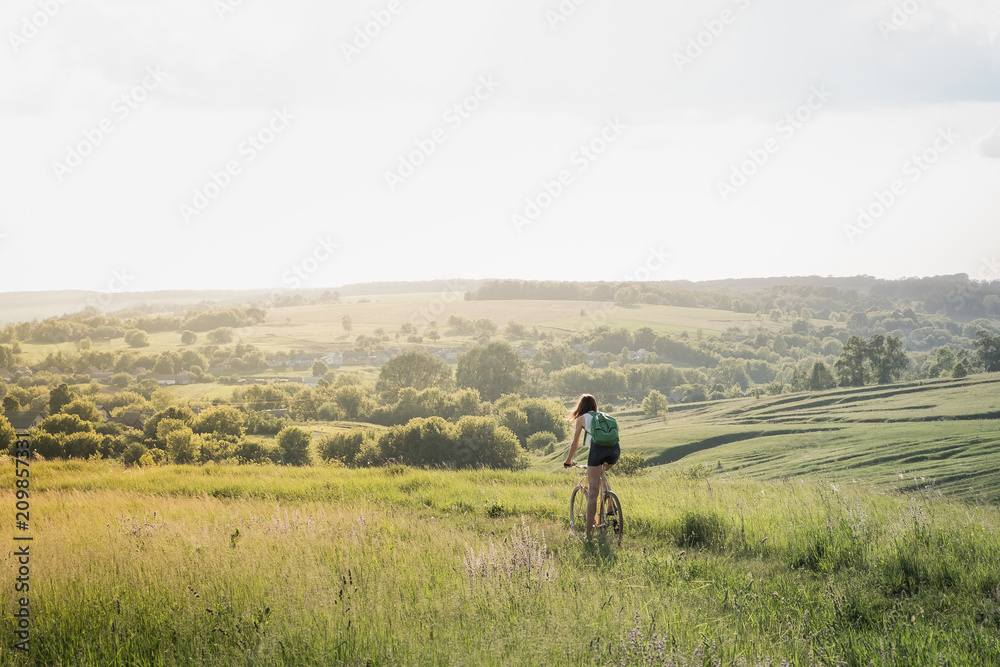 Girl riding a bicycle down the hill in beautiful rural landscape at sunset. Young pretty female person with retro bike standing in a meadow on bright sunny afternoon in summer