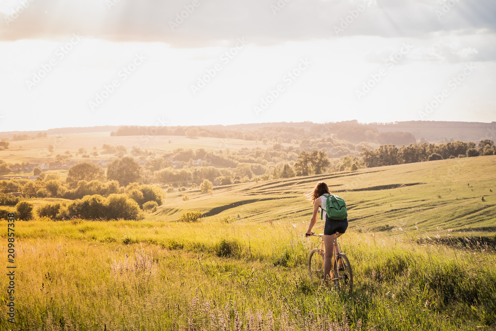 Girl riding a bicycle down the hill in beautiful rural landscape at sunset. Young pretty female person with retro bike standing in a meadow on bright sunny afternoon in summer
