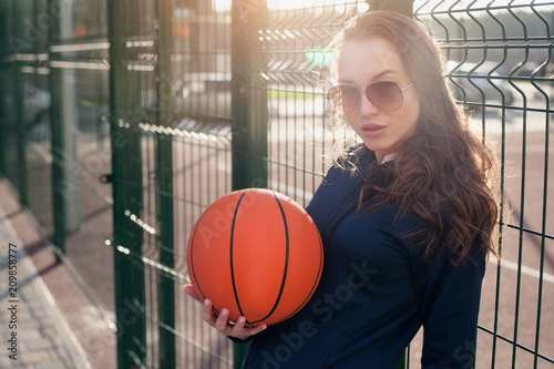 Beautiful girl on a summer evening standing at the Playground and holding a basketball ball. She looks into the camera with a place for your text