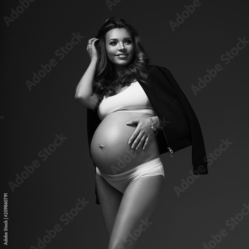 Full body portrait of attractive smiling pregnant brunette woman with slightly curly hair, wearing white sports underwear, trainers and with black jacket draped over shoulders, standing and posing. © Dmitry Tsvetkov