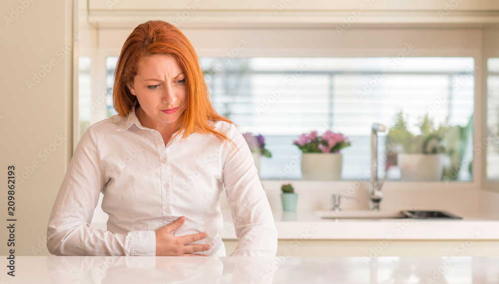 Redhead woman at kitchen with hand on stomach because nausea, painful disease feeling unwell. Ache concept.