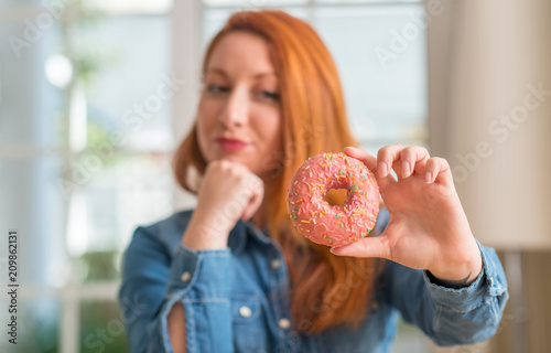 Redhead woman holding donut at home serious face thinking about question, very confused idea