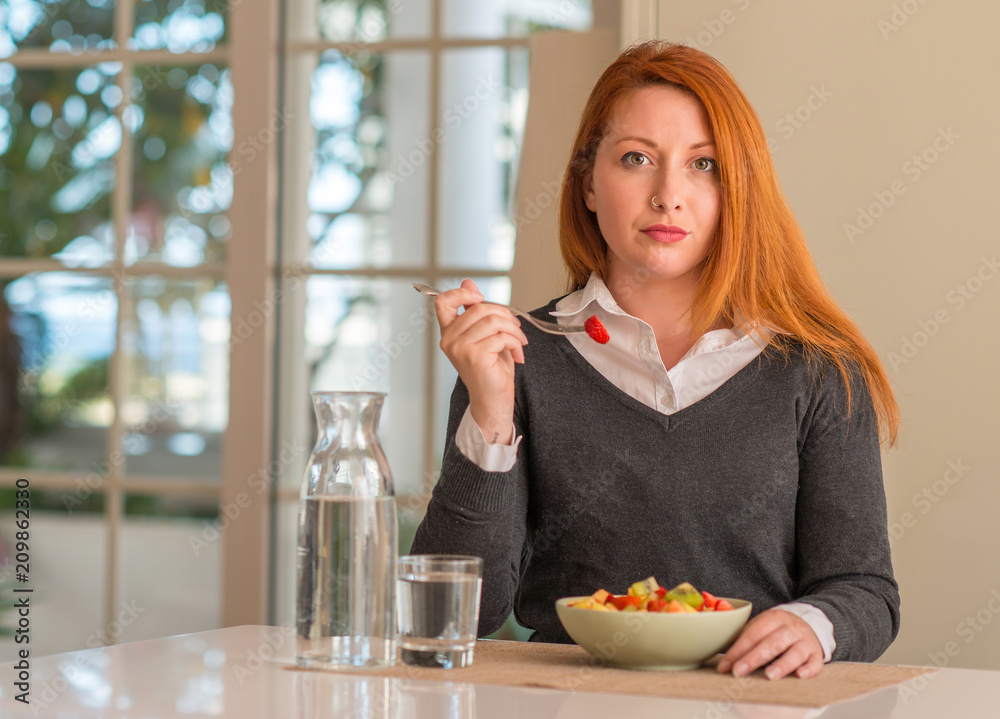 Redhead woman eating fruit bowl, kiwi and strawberry at home with a confident expression on smart face thinking serious