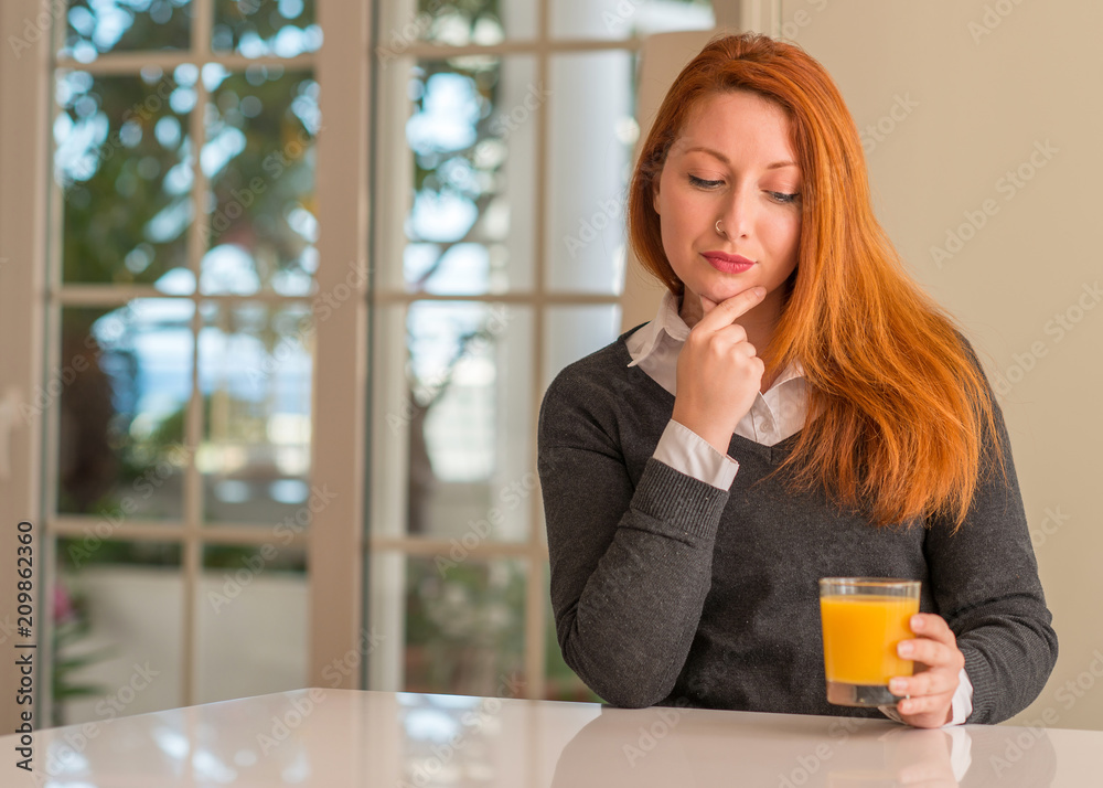 Redhead woman holding orange juice glass at home, serious face thinking about question, very confused idea Redhead woman holding orange juice glass at home, serious face thinking about question, very confused idea