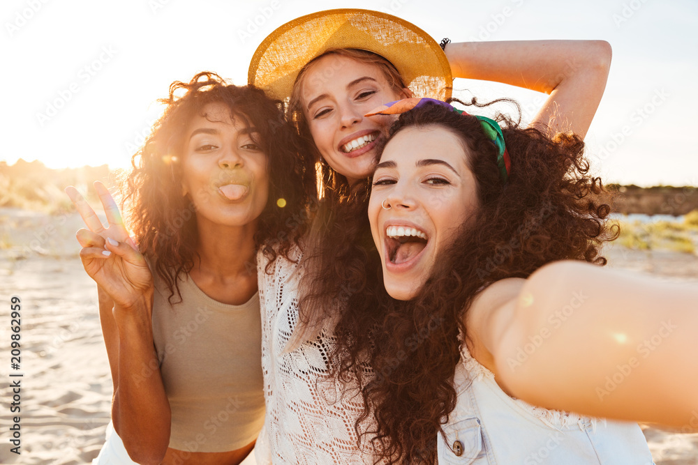 Naklejka premium Three positive multiethnic women 20s in summer clothing smiling at camera, while taking selfie photo during holiday on nature
