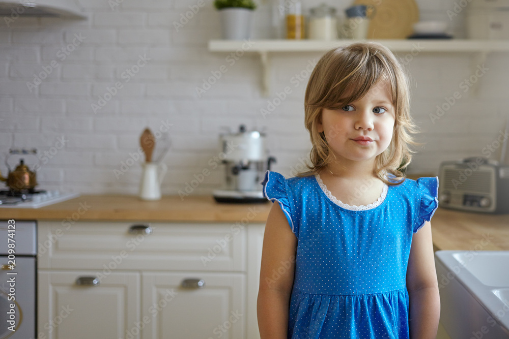 Indoor picture of cute funny little girl with chubby cheeks and fair hair posing in kitchen interior wearing blue dress looking sideways with playful facial expression. People and lifestyle Indoor picture of cute funny little girl with chubby cheeks and fair hair posing in kitchen interior wearing blue dress looking sideways with playful facial expression. People and lifestyle
