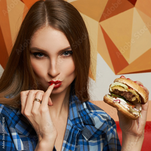Hot attractive long haired female model dressed in blue checkered unbuttoned shirt holding delicious grilled juicy burger and licking her little finger. Sexy beautiful woman at fast food restaurant.